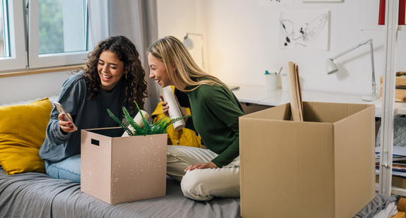 Zwei junge Frauen sitzen mit Umzugskartons und Smartphone auf einem Bett im Wohnheim Zwei junge Frauen sitzen mit Umzugskartons und Smartphone auf einem Bett im Wohnheim